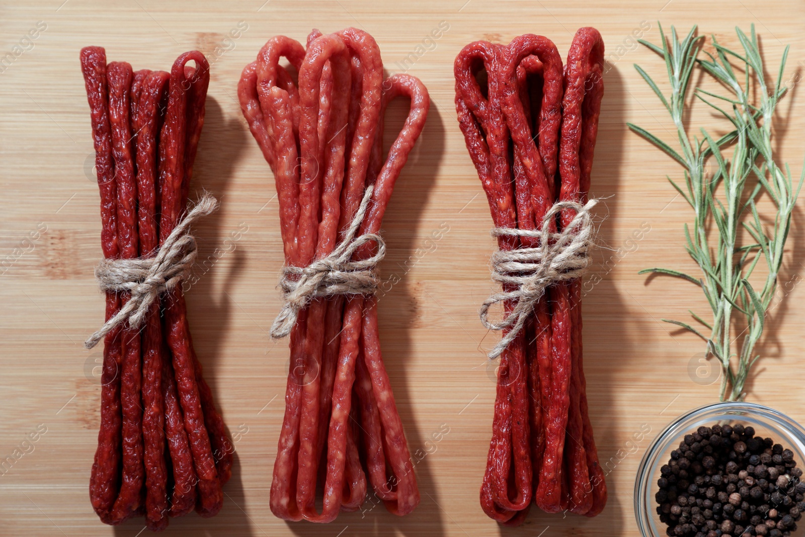 Photo of Bundles of delicious kabanosy with peppercorn and rosemary on wooden table, flat lay