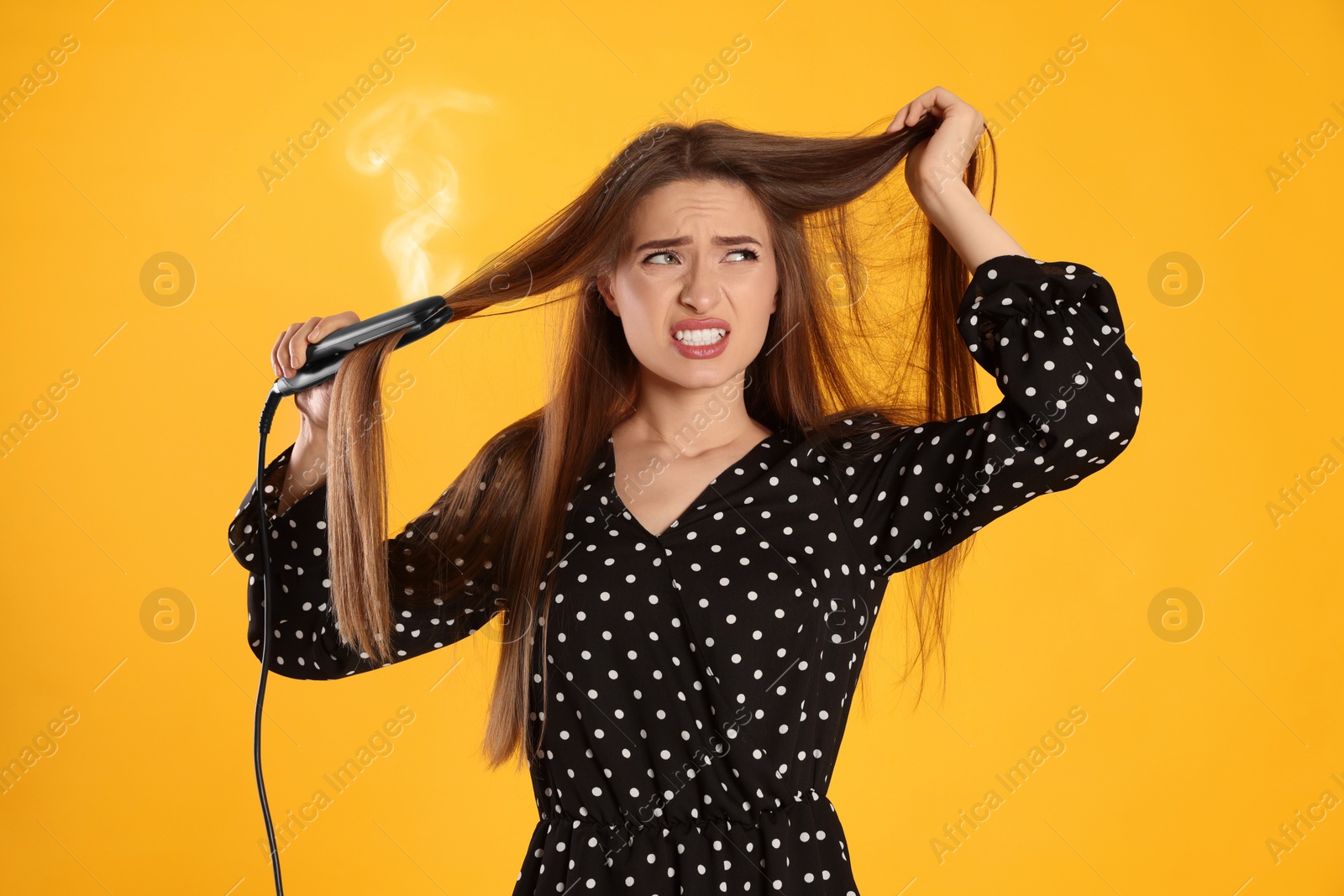 Image of Stressed young woman with flattening iron on yellow background. Hair damage