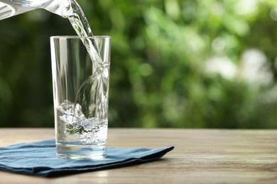 Photo of Pouring water into glass on wooden table outdoors, space for text