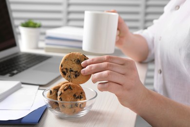 Office worker with cup of drink taking chocolate chip cookie from bowl at workplace, closeup