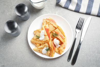 Delicious thin pancakes with salmon and sour cream on grey table, flat lay