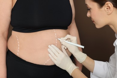 Doctor drawing marks on obese woman's body against brown background, closeup. Weight loss surgery