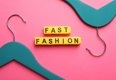 Photo of Yellow plastic cubes with phrase Fast Fashion and hangers on pink background, flat lay