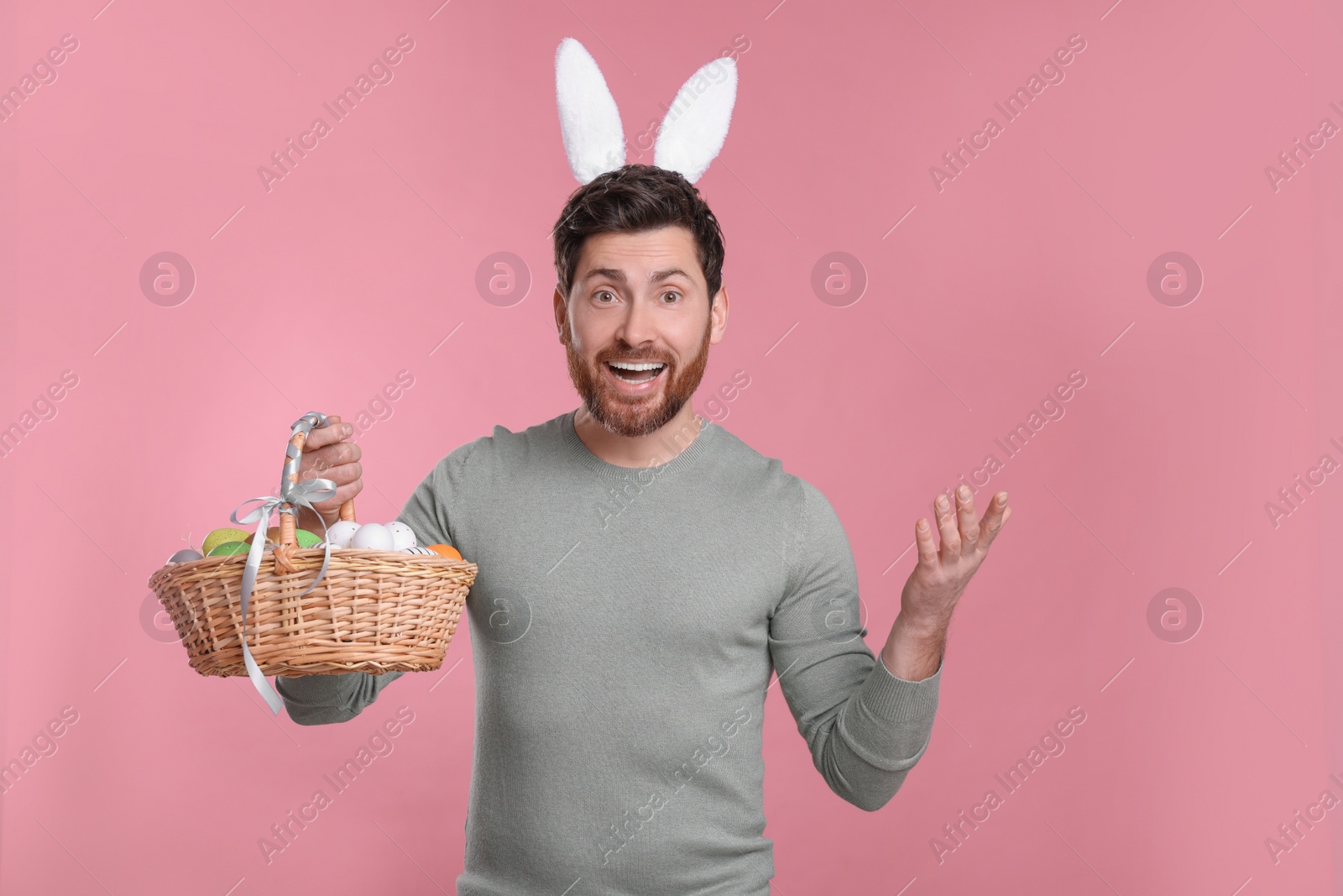 Photo of Portrait of happy man in cute bunny ears headband holding wicker basket with Easter eggs on pink background