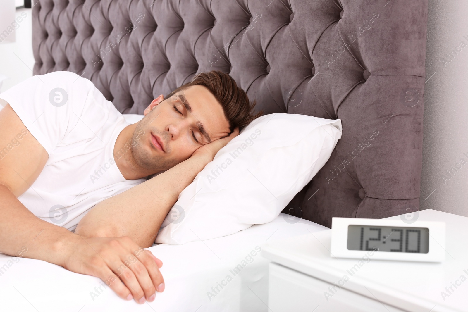 Photo of Young man sleeping in bed and electronic alarm clock on nightstand at home