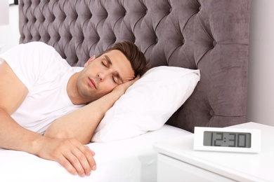 Young man sleeping in bed and electronic alarm clock on nightstand at home