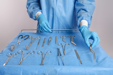 Photo of Doctor holding medical forceps near table with different surgical instruments on light background, closeup