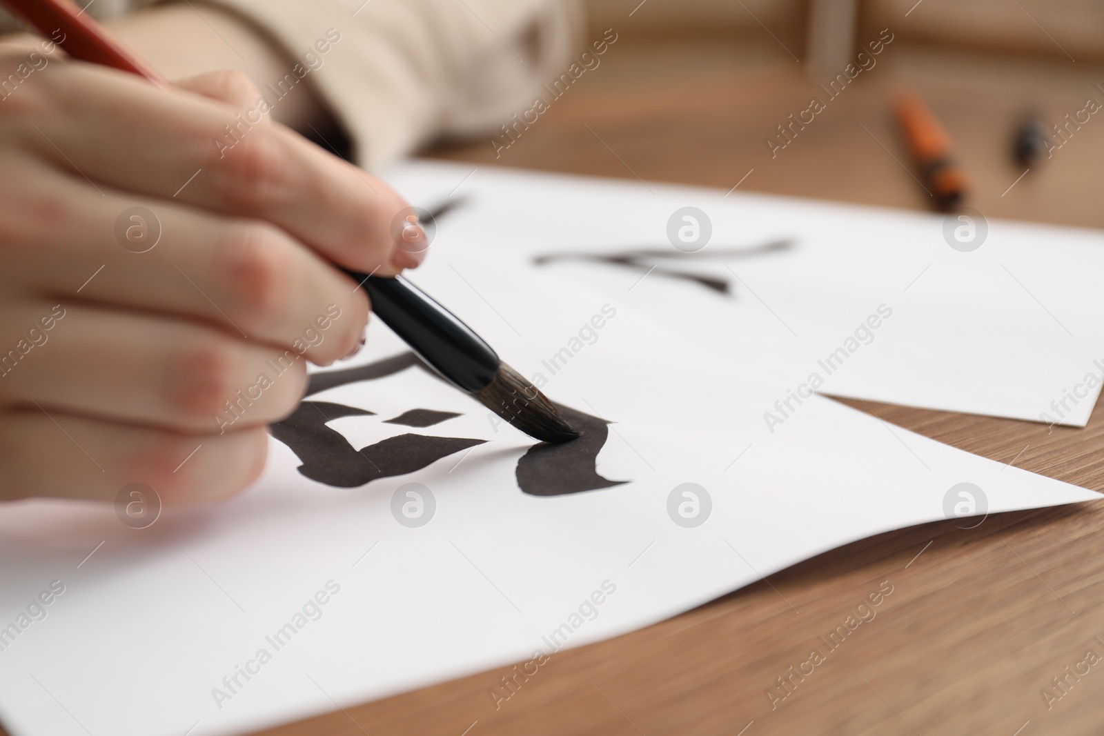 Photo of Calligraphy. Woman with brush writing hieroglyphs on paper at wooden table, closeup