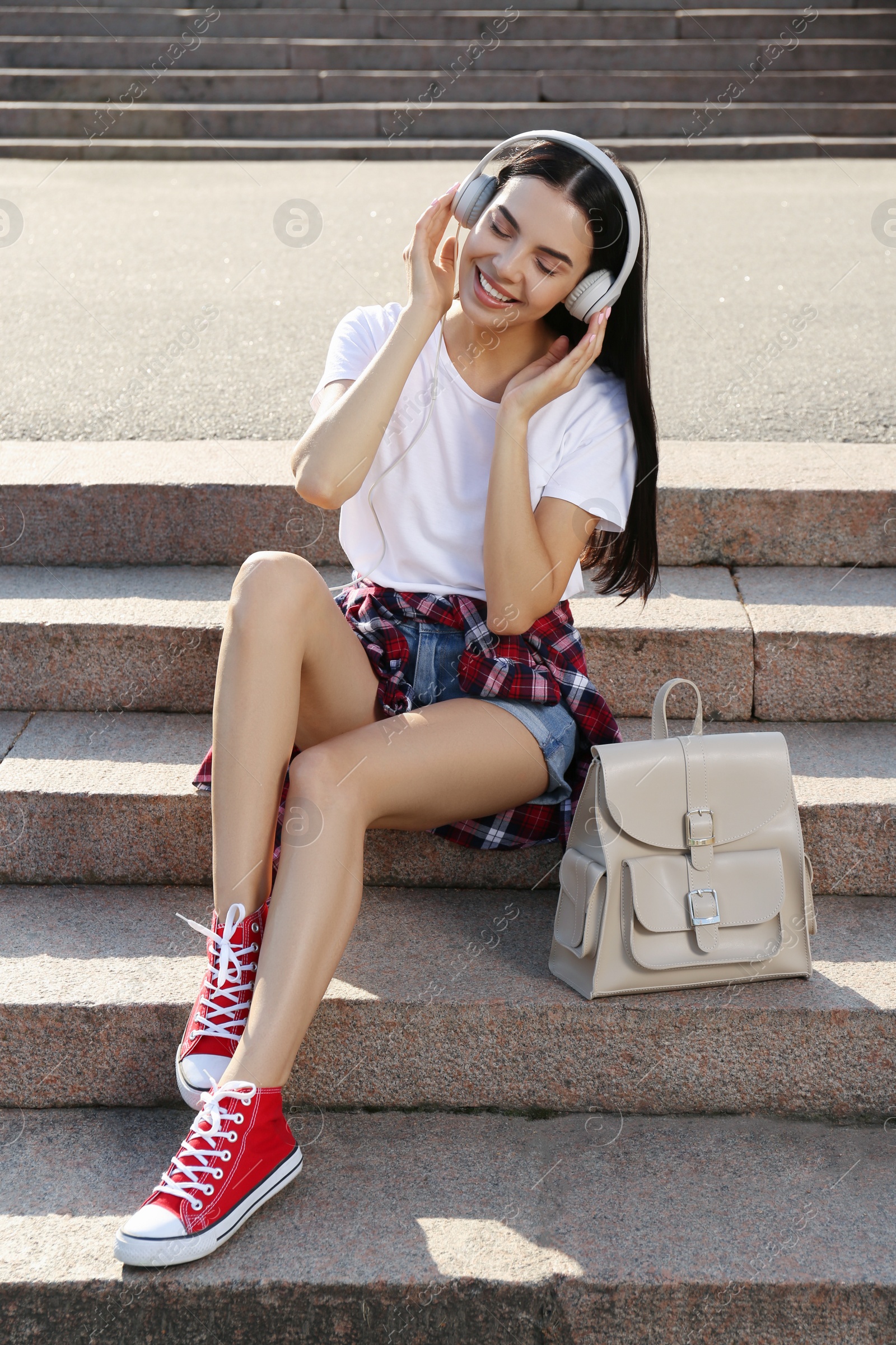 Photo of Beautiful young woman with stylish beige backpack and headphones on stairs outdoors