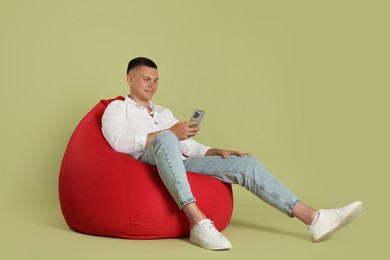 Handsome man with smartphone on red bean bag chair against green background