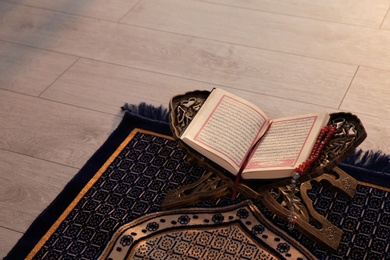 Photo of Rehal with open Quran and Muslim prayer beads on rug indoors. Space for text