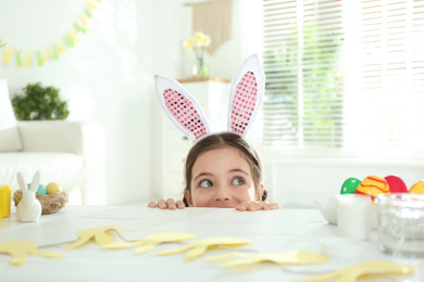 Photo of Cute little girl wearing bunny ears headband at table with Easter eggs, indoors