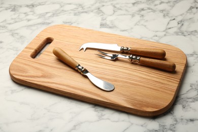 Wooden cutting board and cutlery on white marble table, closeup