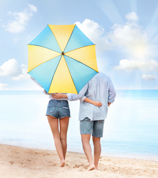 Image of Happy young couple with umbrella for sun protection walking on beach near sea, back view