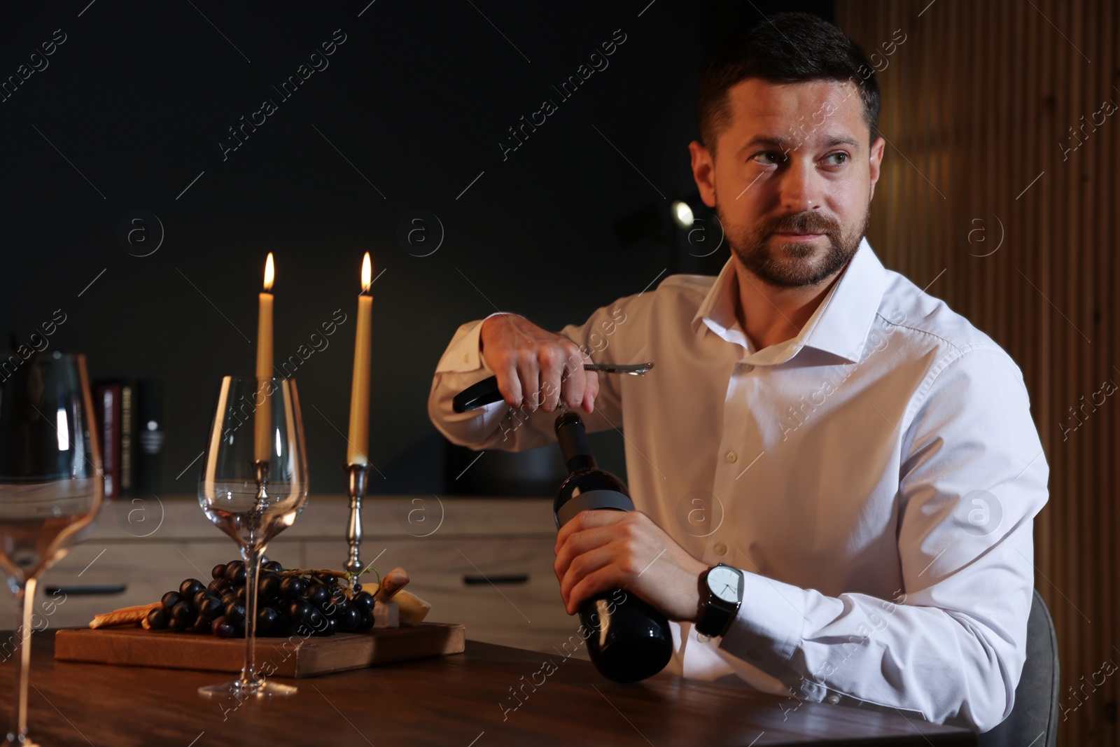 Photo of Romantic dinner. Man opening wine bottle with corkscrew at table indoors