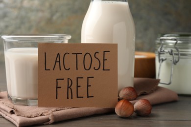 Photo of Milk in glassware, card with phrase Lactose free and hazelnuts on wooden table, closeup
