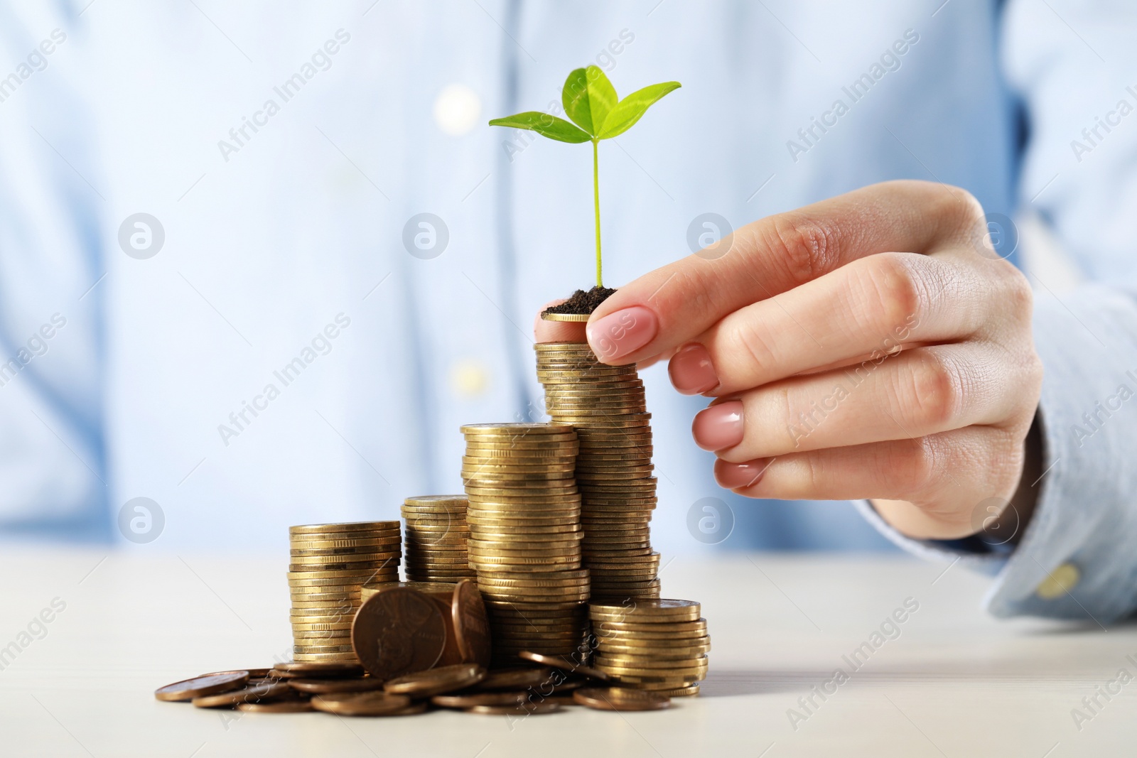 Photo of Woman putting coin with green sprout onto stack at white table, closeup. Investment concept