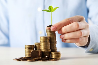Photo of Woman putting coin with green sprout onto stack at white table, closeup. Investment concept