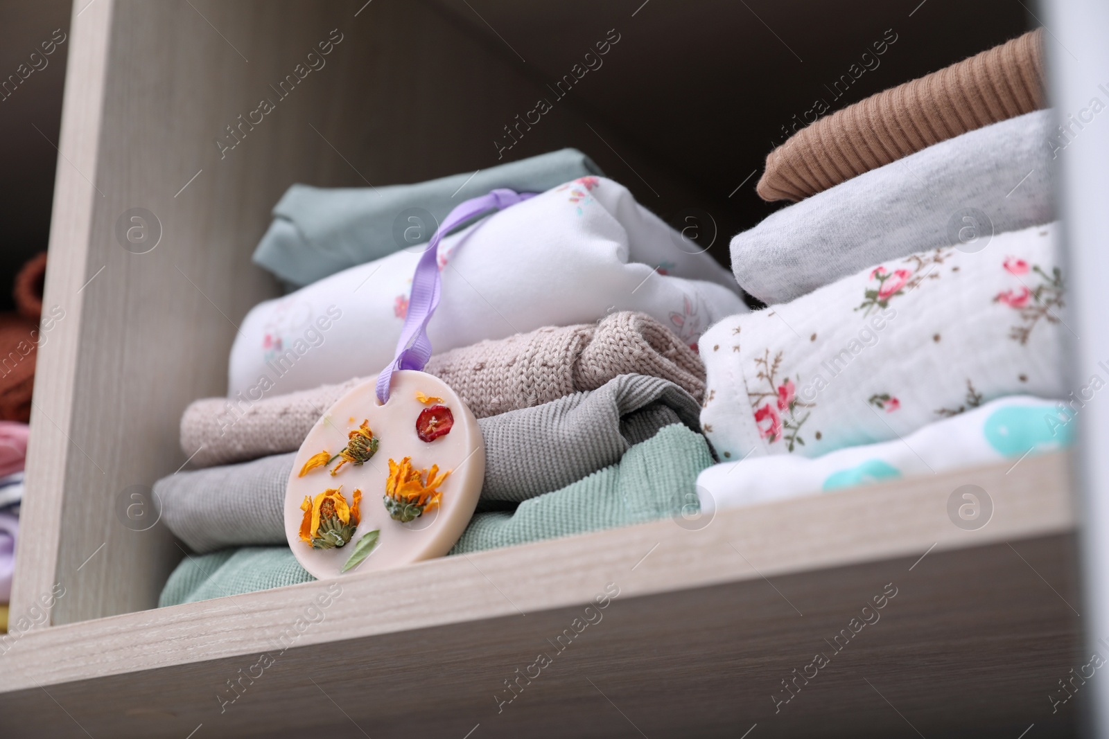 Photo of Beautiful scented wax sachet near stack of clothes on shelf in wardrobe, closeup