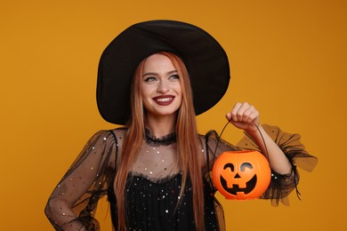 Photo of Happy young woman in scary witch costume with pumpkin bucket on orange background. Halloween celebration