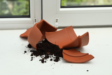 Broken terracotta flower pot with soil on white windowsill indoors