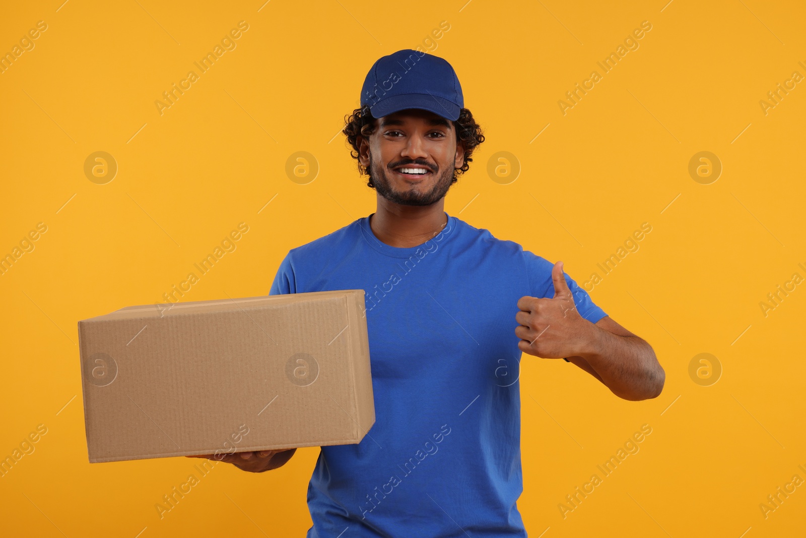 Photo of Happy young courier with parcel showing thumb up on orange background