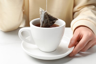 Woman putting tea bag in cup with hot water at white table, closeup