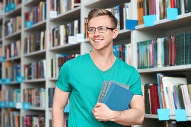 Photo of Young man with books near shelving unit in library