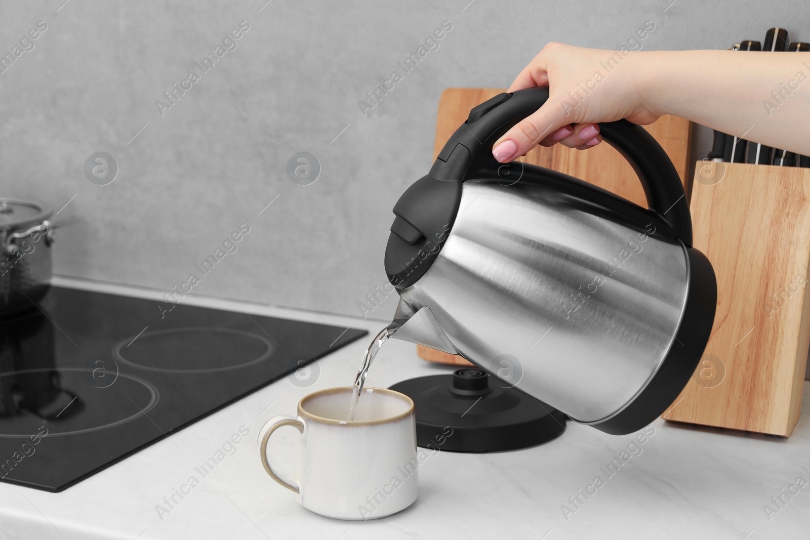 Photo of Woman pouring hot water from electric kettle into cup in kitchen, closeup