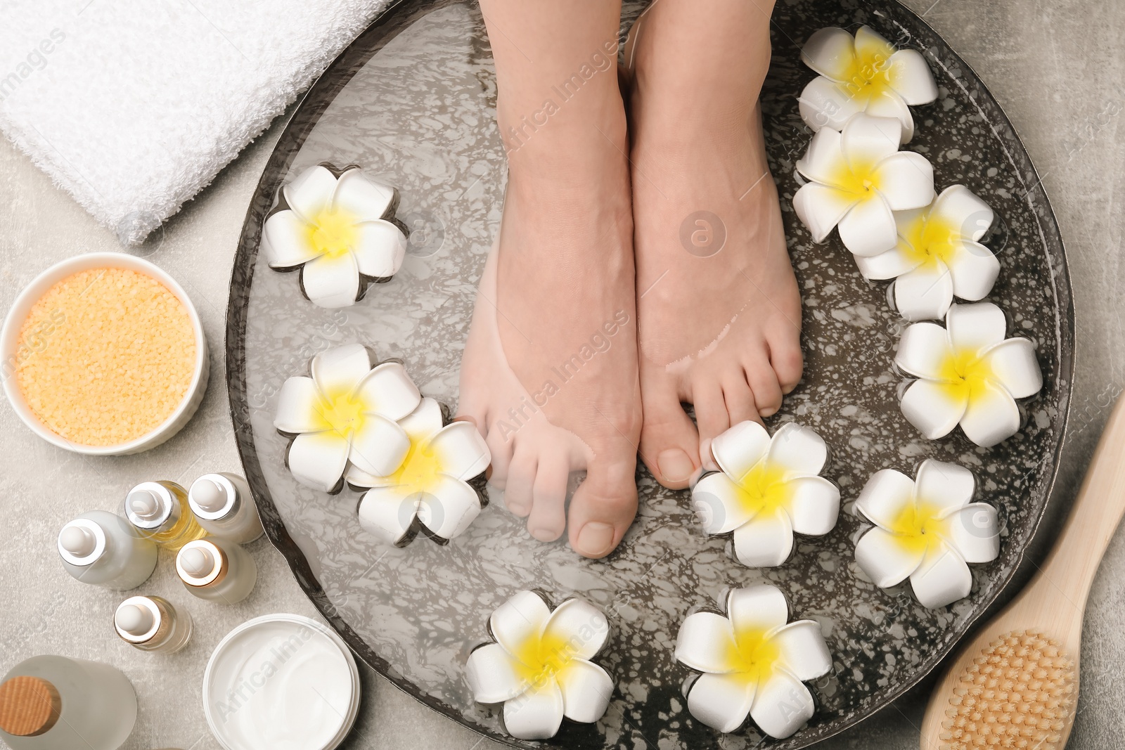 Photo of Woman soaking her feet in bowl with water and flowers on light grey floor, top view. Spa treatment