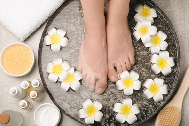 Photo of Woman soaking her feet in bowl with water and flowers on light grey floor, top view. Spa treatment