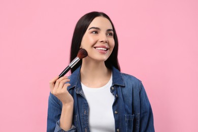 Beautiful woman applying makeup with brush on pink background