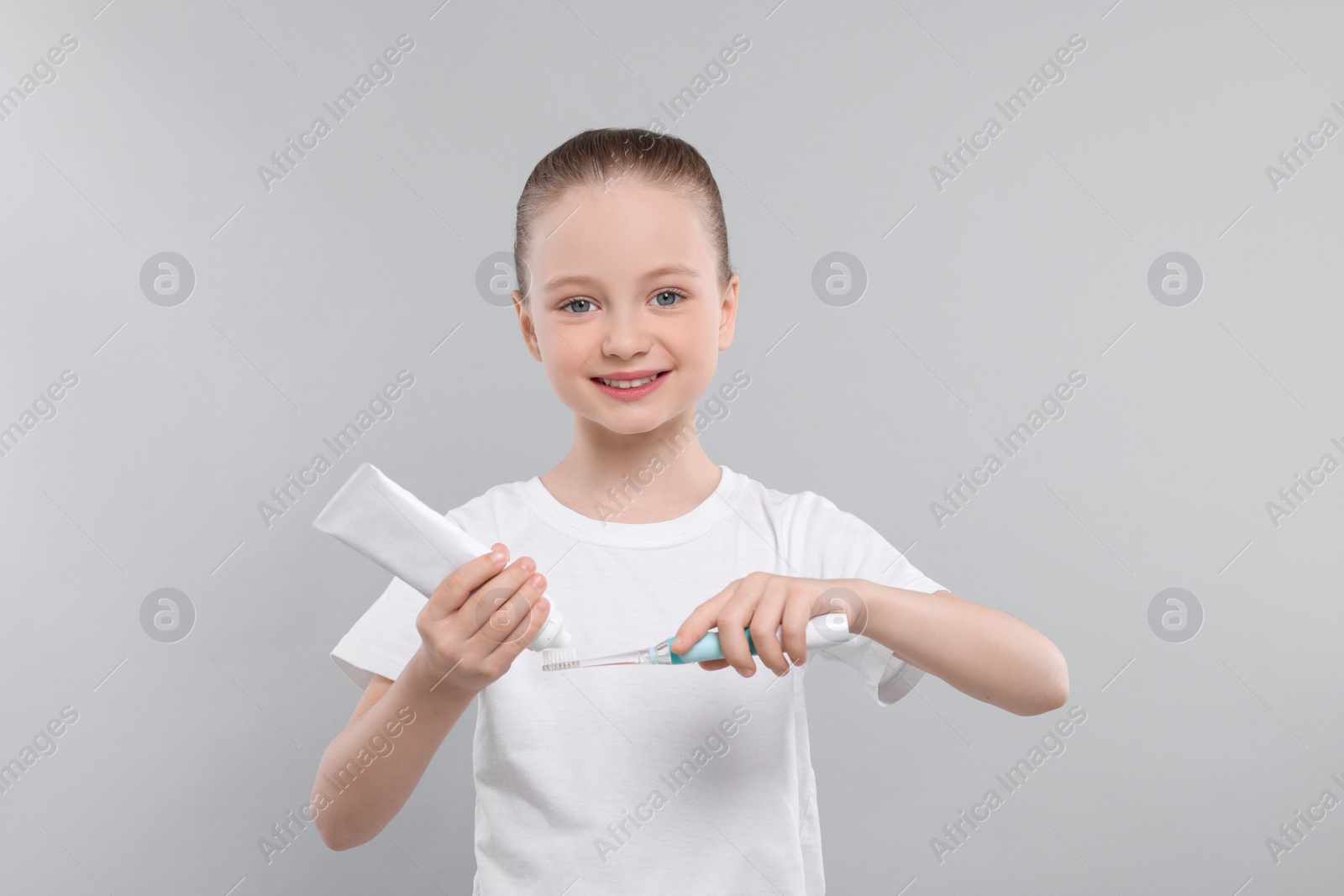 Photo of Happy girl squeezing toothpaste from tube onto electric toothbrush on light grey background