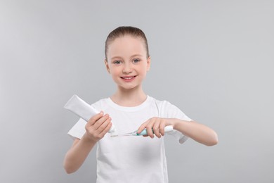 Happy girl squeezing toothpaste from tube onto electric toothbrush on light grey background