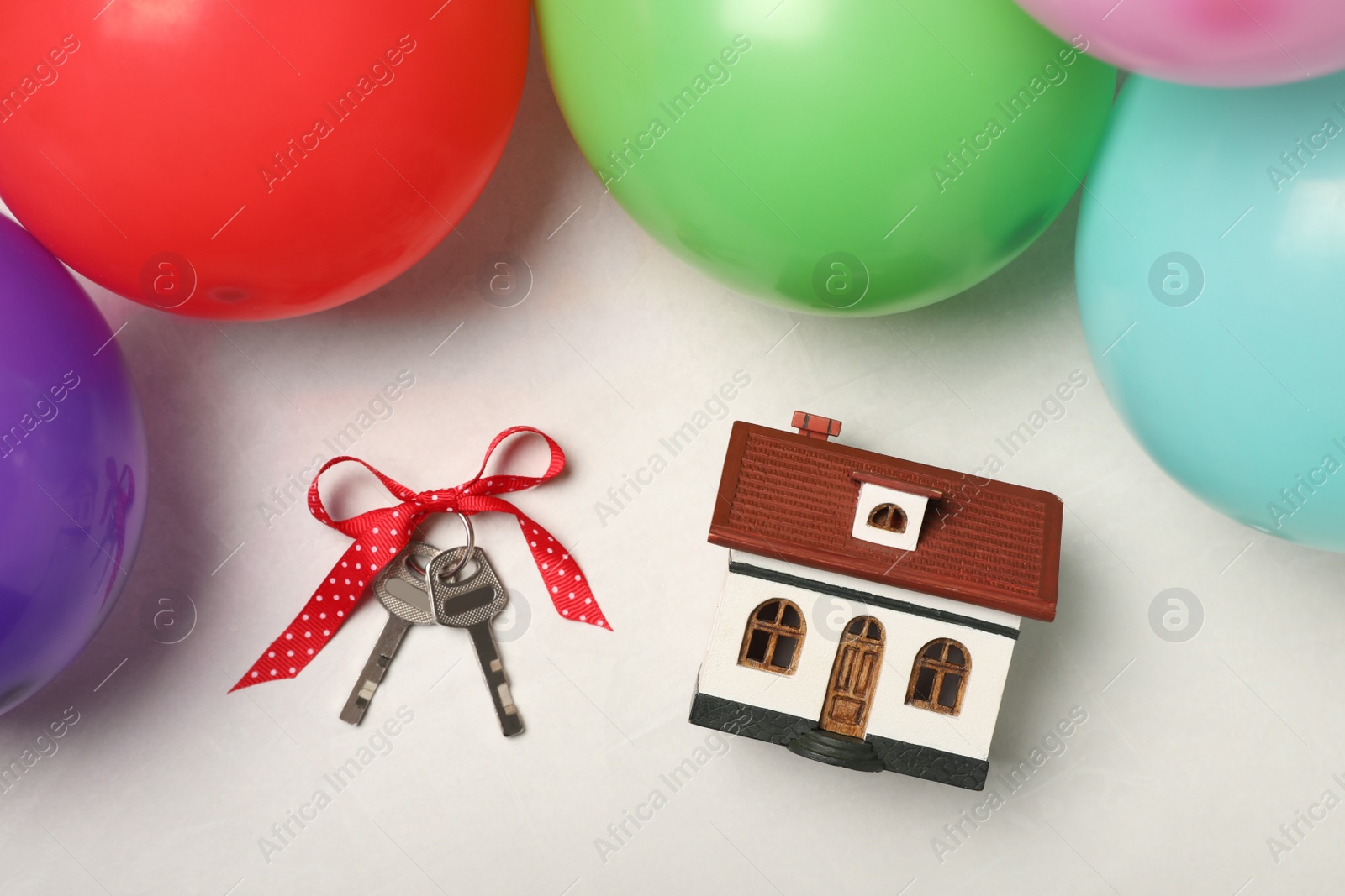 Photo of Wooden house model, keys with bow and balloons on white table, flat lay. Housewarming party