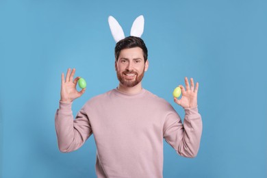 Happy man in cute bunny ears headband holding Easter eggs on light blue background