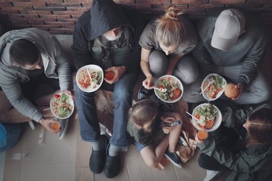 Image of Poor people with plates of food sitting at wall indoors, view from above