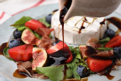 Pouring balsamic vinegar onto fresh salad with brie cheese, figs and berries on plate, closeup