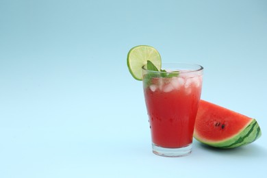 Photo of Glass of delicious drink with lime, ice cubes and cut fresh watermelon on light blue background, space for text