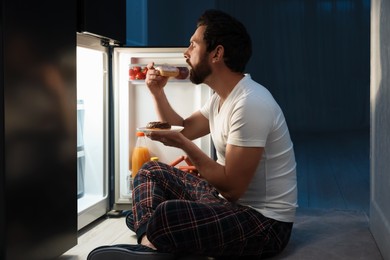 Photo of Man eating donuts near refrigerator in kitchen at night. Bad habit