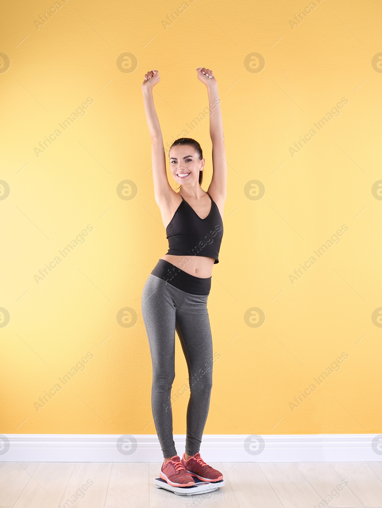 Photo of Happy young woman measuring her weight using scales near color wall. Weight loss motivation