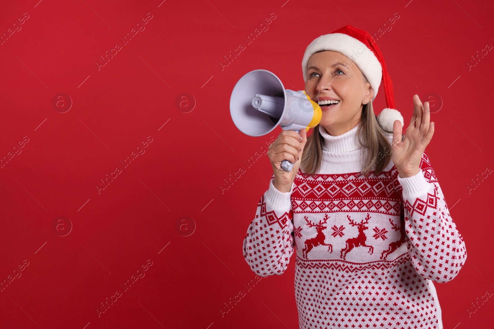 Photo of Senior woman in Christmas sweater and Santa hat shouting in megaphone on red background. Space for text