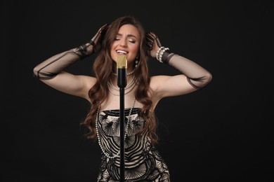 Beautiful young woman in stylish dress with microphone singing on black background