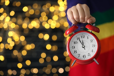 Woman holding alarm clock against blurred lights, closeup with space for text. New Year countdown