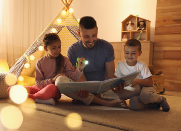 Father and children with flashlight reading book at home