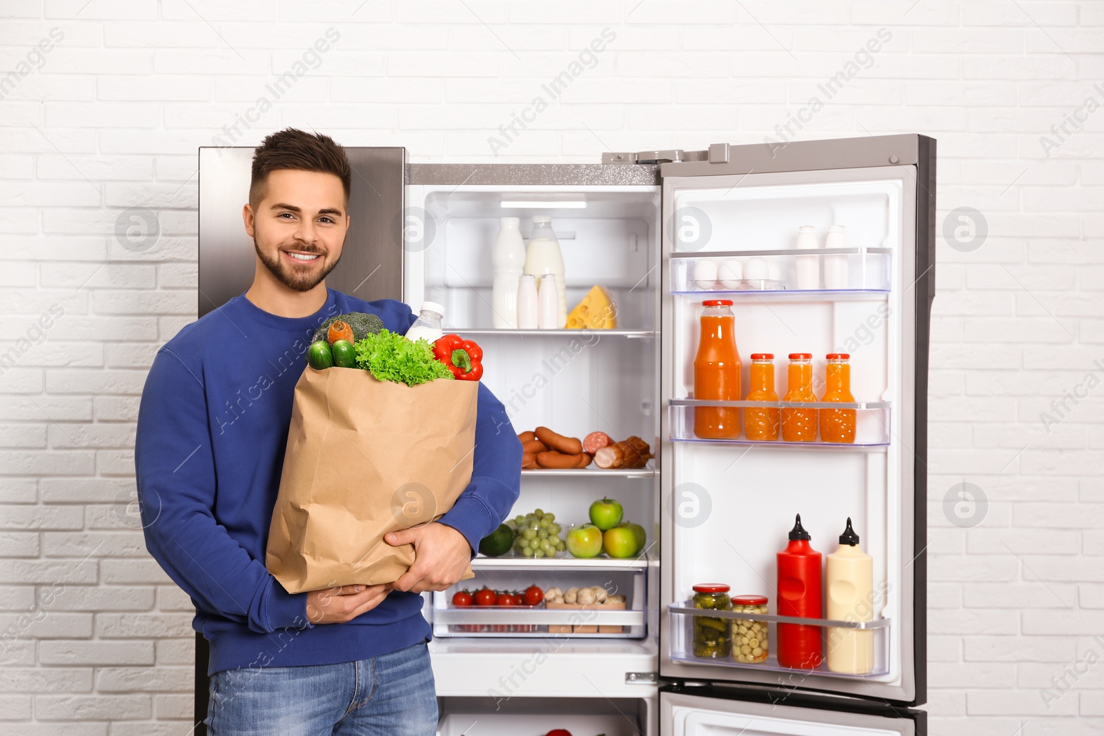 Photo of Young man with paper bag full of products near open refrigerator indoors