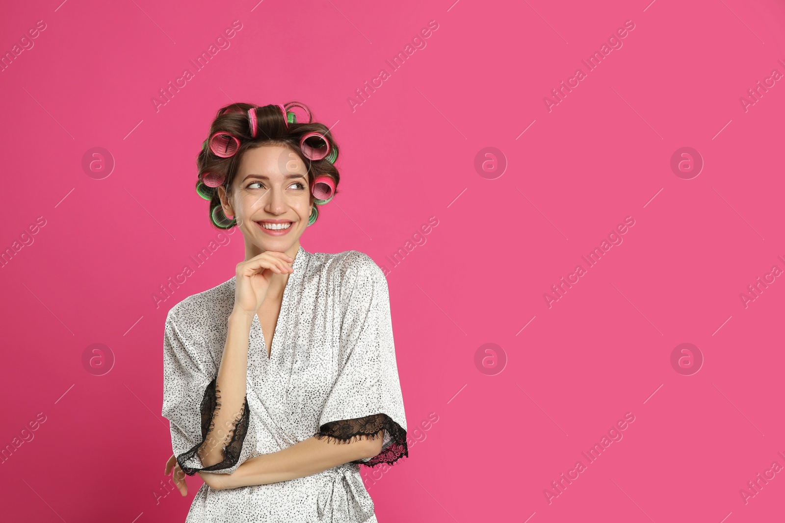 Photo of Beautiful young woman in silk bathrobe with hair curlers on pink background. Space for text