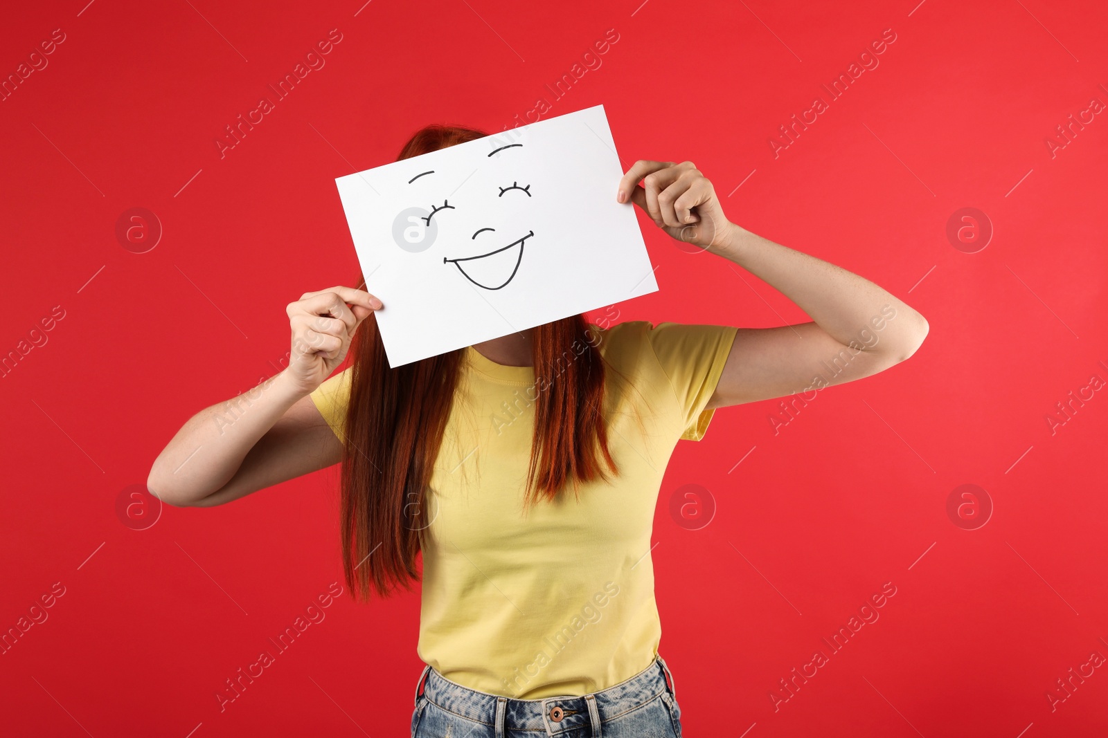 Photo of Woman hiding behind sheet of paper with happy face on red background