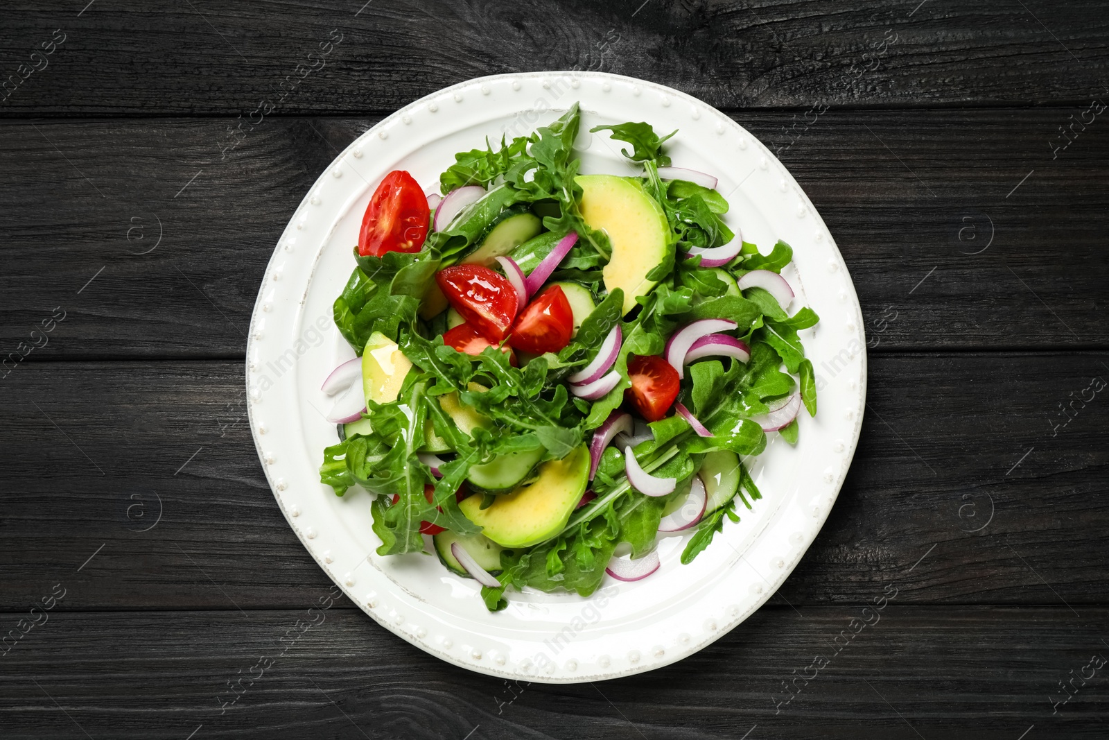 Photo of Delicious salad with avocado, arugula and tomatoes on black wooden table, top view