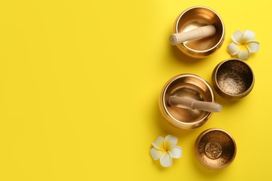 Photo of Golden singing bowls, mallets and flowers on yellow background, flat lay. Space for text